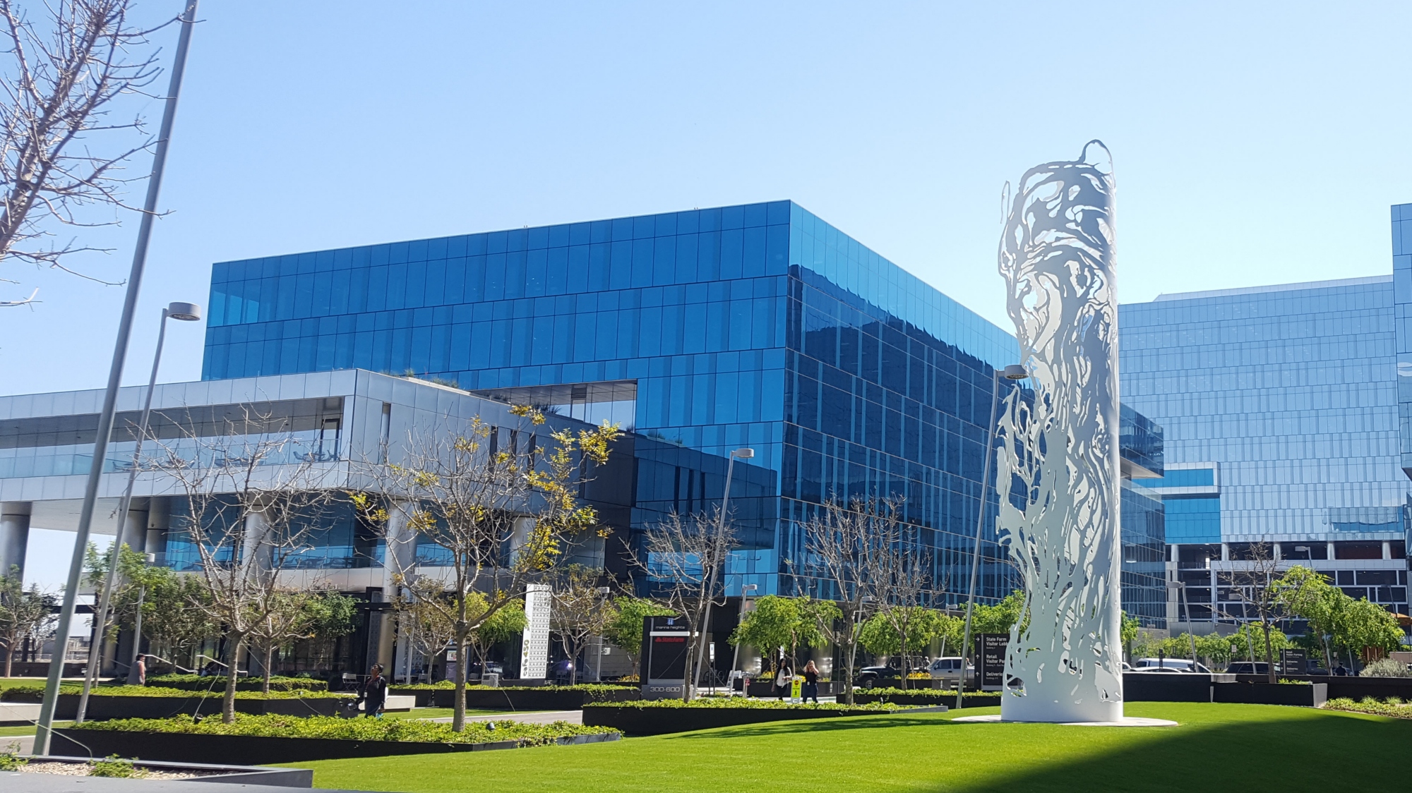 A large glass building sits in the background. In the foreground is a grassy lawn with trees and a tall white sculpture. The sculpture dwarfs the trees and appears to be made out of a thin metal. The metal has holes cut into it that make the sculpture look like a wispy, light stream of water.