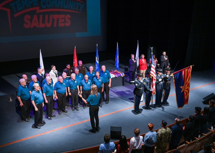 A group of singers sings on stage alongside Color Guard and elected officials with hands over hearts singing the national anthem