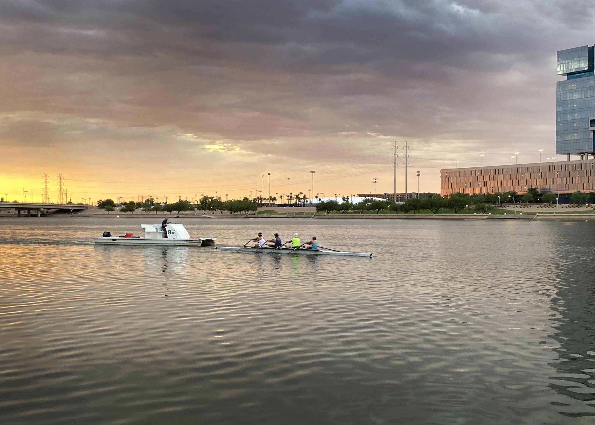 A rowing team of four people rows on Tempe Town Lake in the sliver of sunlight peeking up above the horizon.